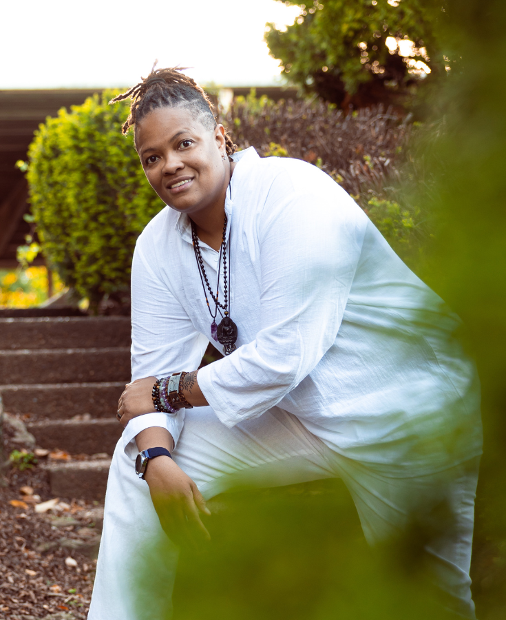 Meditation teacher Shawn dressed in white linen with shrubs and bushes in the foreground and background leaning on her knee off the path of steps that are behind her.